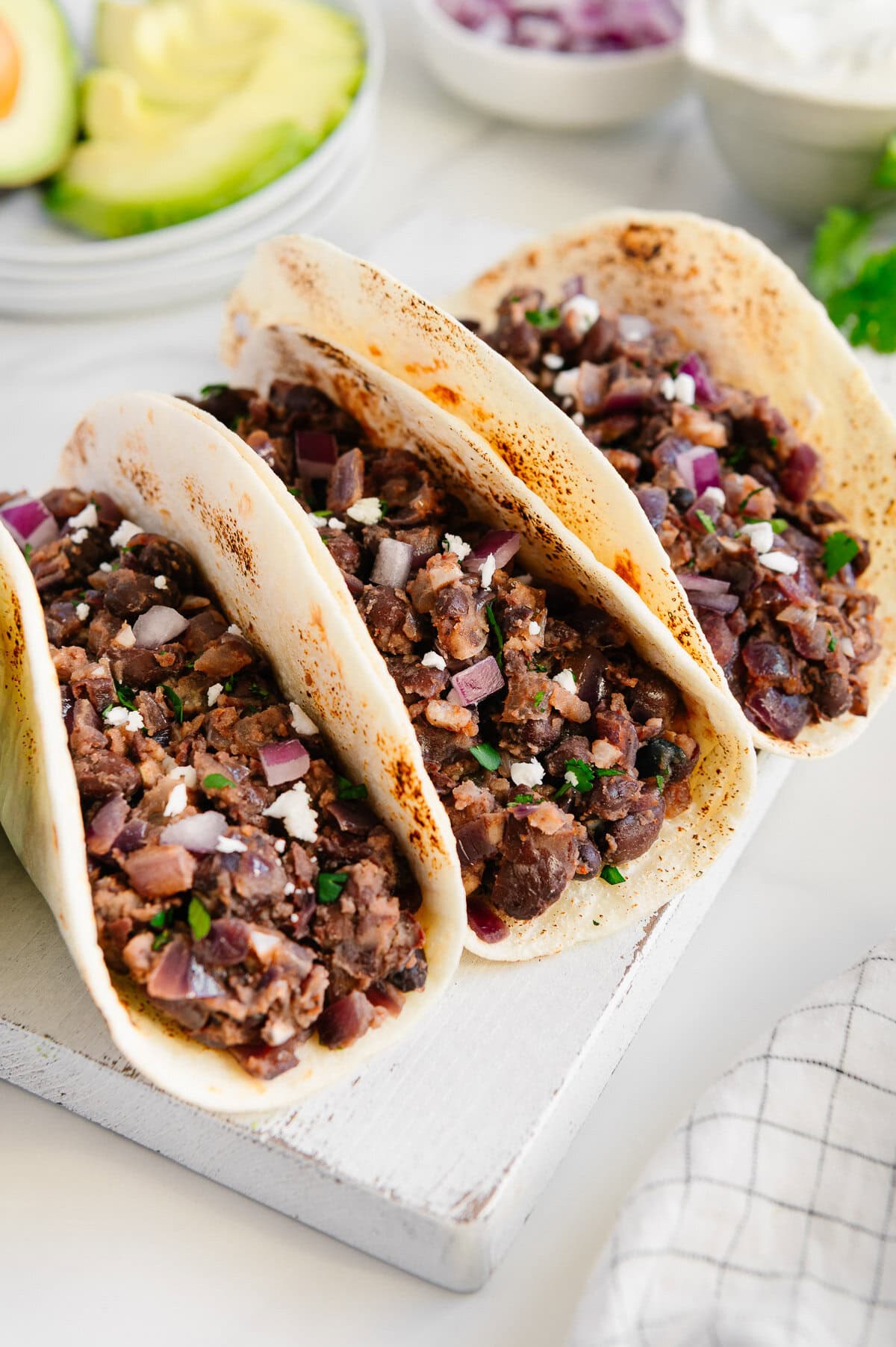 Three soft tacos filled with a black bean and vegetable mixture, topped with crumbled cheese, are arranged on a white board. Sliced avocado, onion, and a bowl of sour cream are in the background.