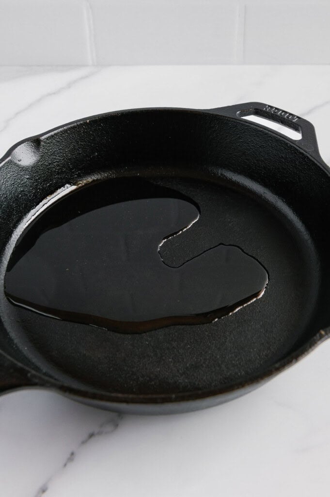 A cast iron skillet sits on a white marble countertop with a small amount of oil spread across its surface.