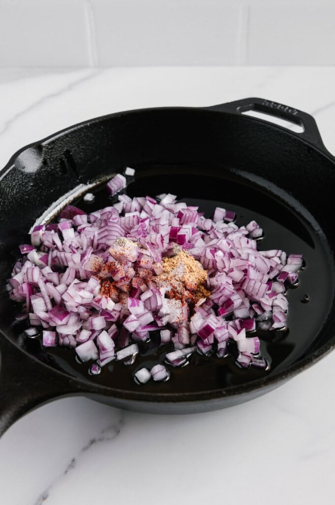 Diced red onions and spices in a black cast iron skillet with oil, placed on a white marble countertop.