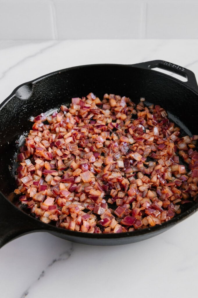 A cast iron skillet filled with sautéed diced red onions sits on a white marble countertop, with the onions appearing caramelized and glossy.