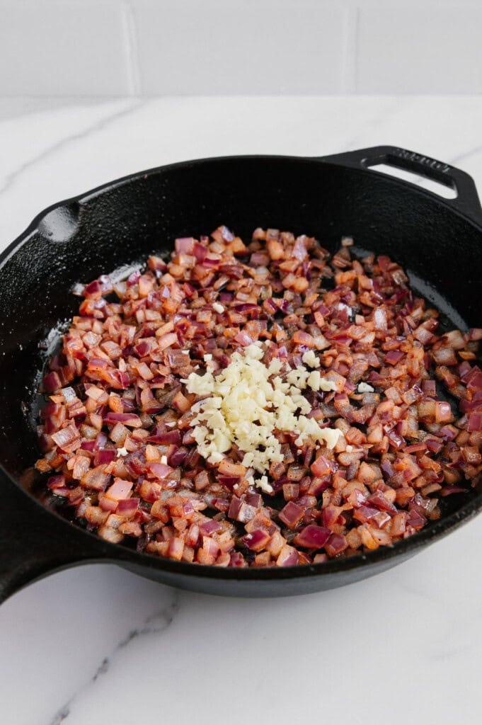 Diced red onions sautéing in a black cast iron skillet, with freshly minced garlic added on top, on a white marble countertop.