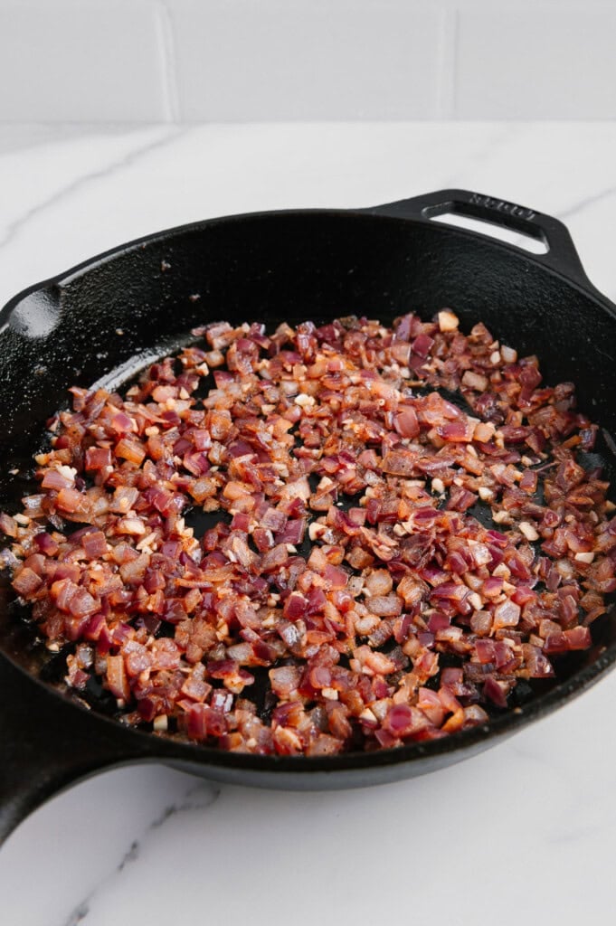 Chopped red onions sautéing in a black cast iron skillet on a white marble countertop, beginning to soften and caramelize.