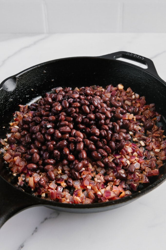 A cast iron skillet on a marble countertop filled with sautéed diced red onions and topped with a mound of black beans.