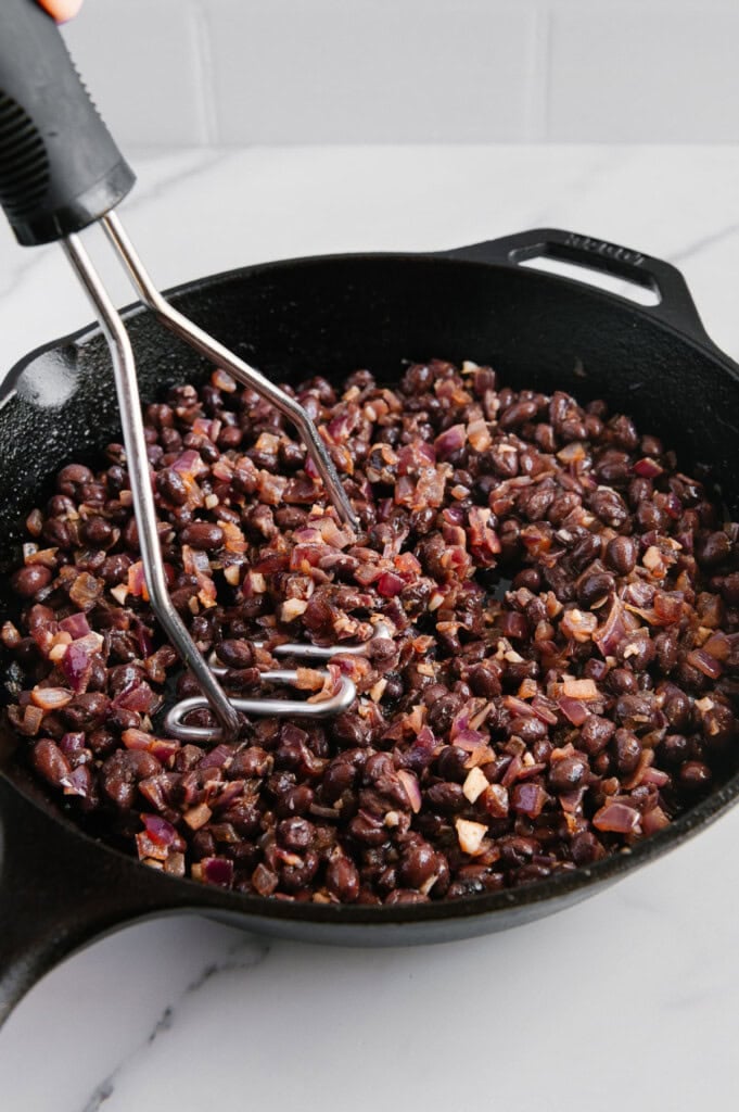 A hand holding a potato masher is mashing black beans and chopped red onions in a black cast iron skillet on a white countertop.