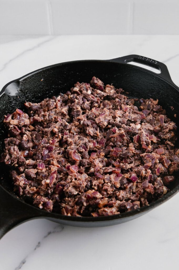 A black cast iron skillet filled with mashed black beans and pieces of diced red onion sits on a white marble countertop against a white tile backsplash.