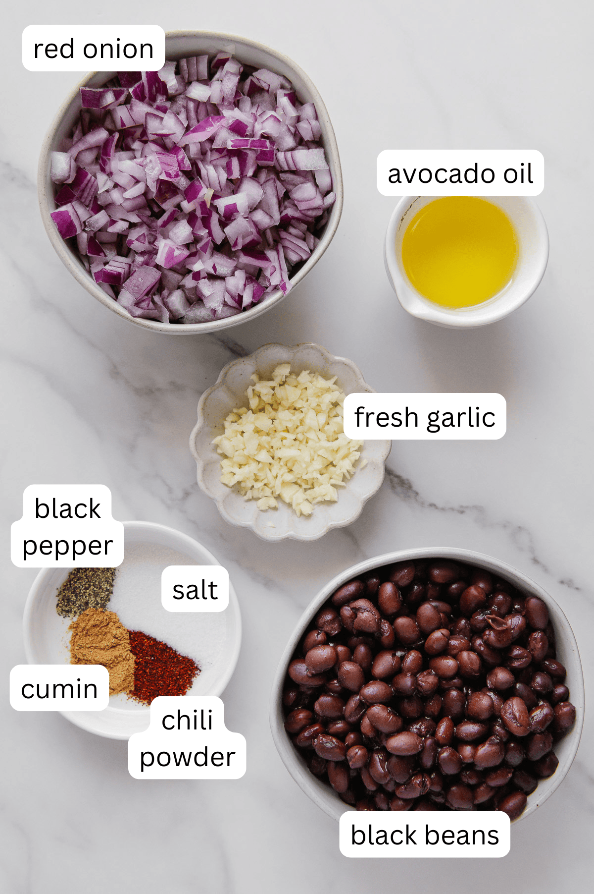 Overhead view of bowls with chopped red onion, mashed black beans, diced fresh garlic, avocado oil, and a dish of salt, black pepper, cumin, and chili powder-perfect toppings for burritos or tacos-labeled on a white marble surface.