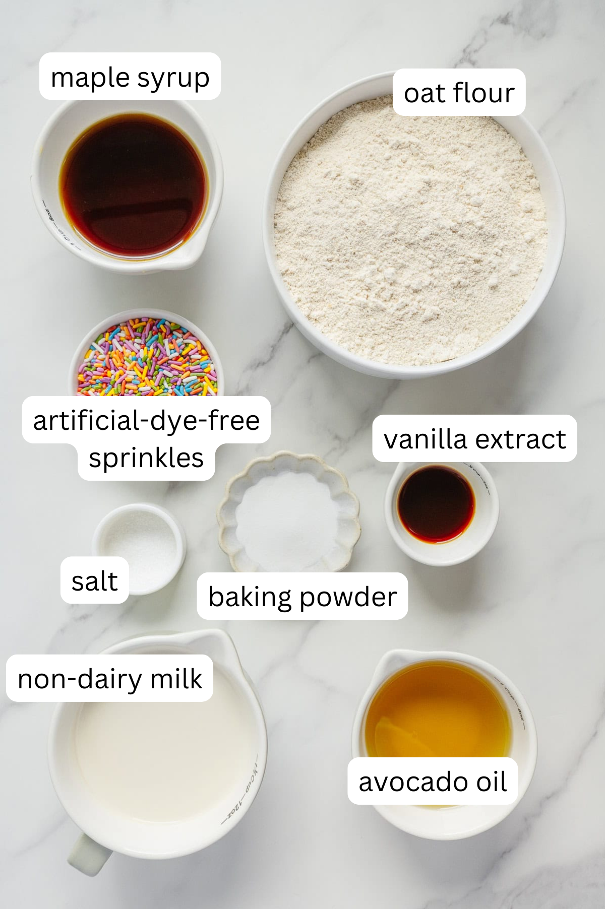 Ingredients for baking are arranged on a white surface, labeled: maple syrup, oat flour, artificial-dye-free sprinkles, vanilla extract, salt, baking powder, non-dairy milk, and avocado oil.