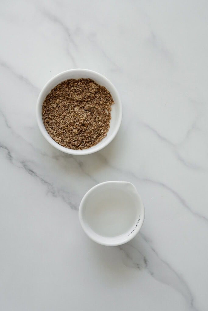 A small white bowl filled with ground flaxseed sits above a white bowl containing clear water, both placed on a white marble surface.