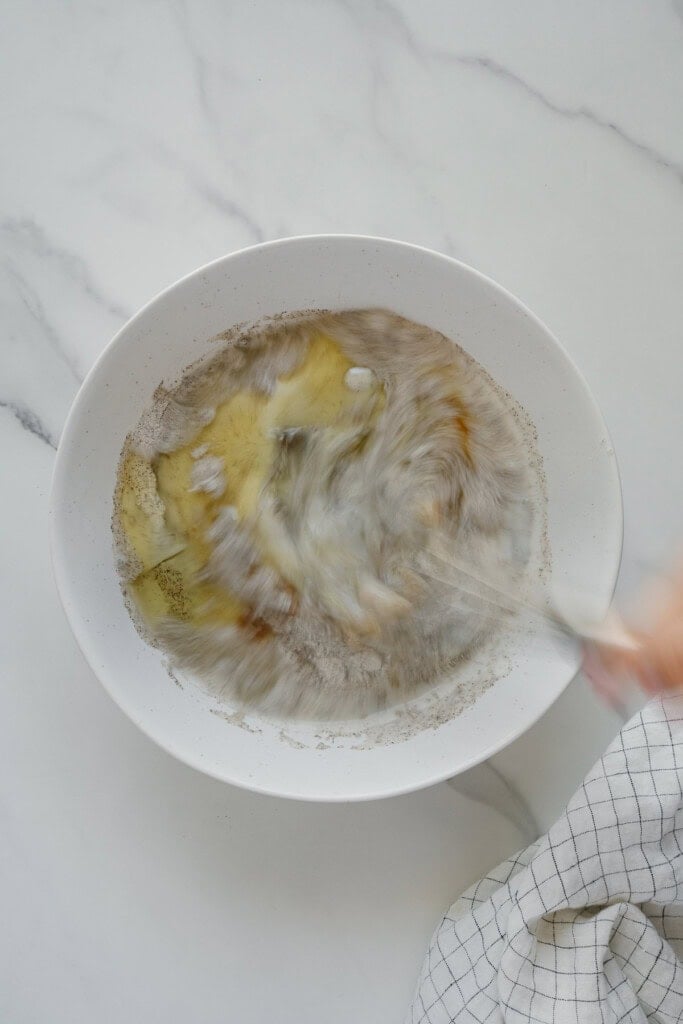 A hand whisks wet and dry ingredients together in a white bowl on a marble countertop, with a checkered kitchen towel nearby.