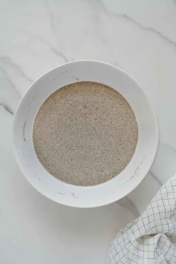 A white bowl filled with a light brown, speckled batter sits on a white marble surface next to a white cloth with a black grid pattern.