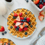 A plate of golden waffles topped with fresh strawberries, raspberries, and blueberries sits on a white surface, surrounded by a small pitcher of syrup, a bowl of mixed berries, cutlery, and a checkered napkin.