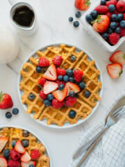 A plate of golden waffles topped with fresh strawberries, raspberries, and blueberries sits on a white surface, surrounded by a small pitcher of syrup, a bowl of mixed berries, cutlery, and a checkered napkin.