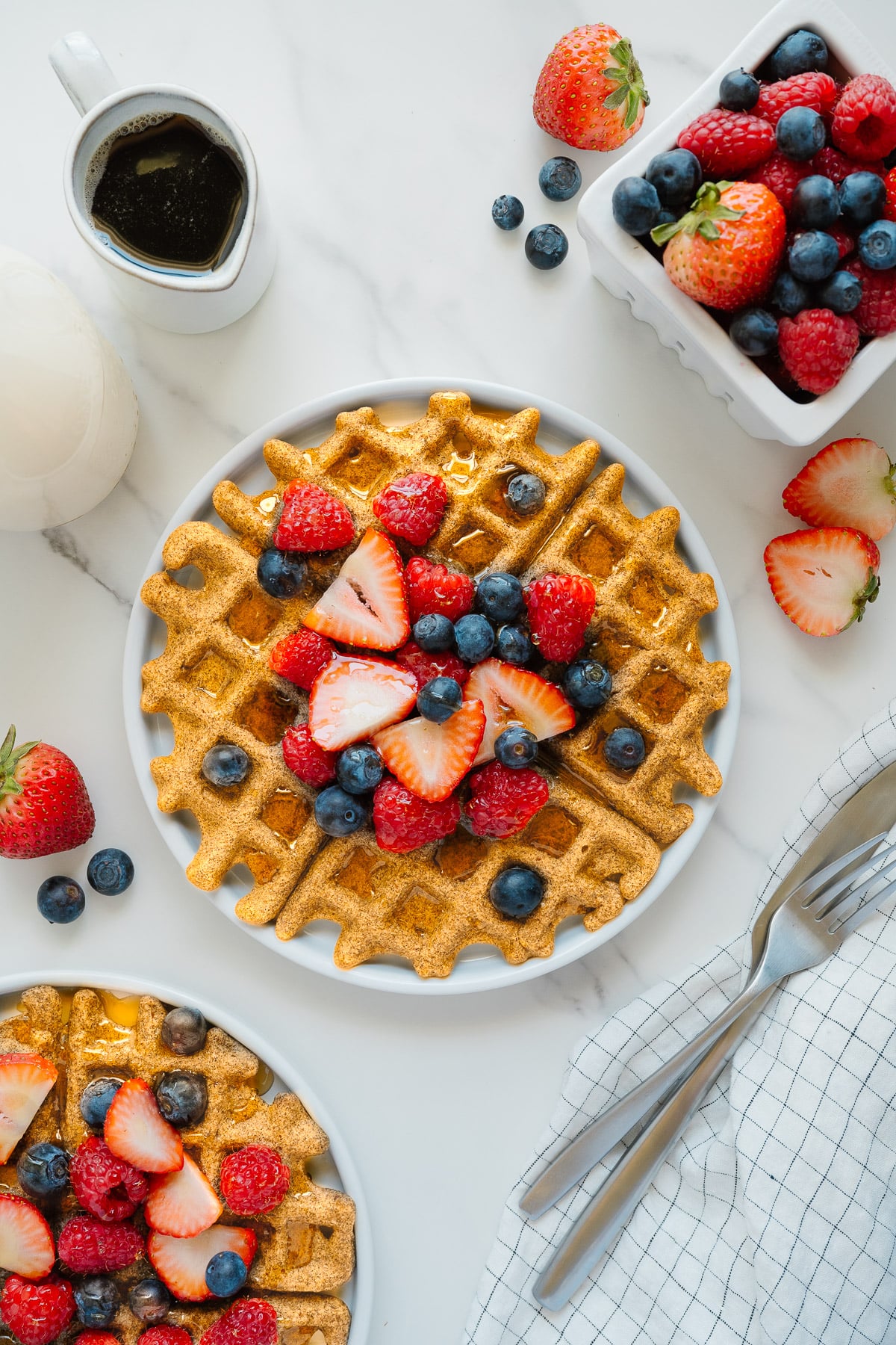 A plate of golden waffles topped with fresh strawberries, raspberries, and blueberries sits on a white surface, surrounded by a small pitcher of syrup, a bowl of mixed berries, cutlery, and a checkered napkin.