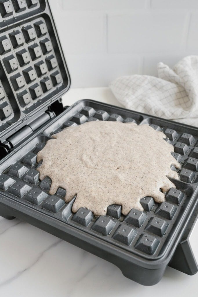 An open waffle maker with uncooked vegan buckwheat waffle batter spread over the grid, ready to be cooked. A white and gray checkered cloth is in the background on a white countertop.