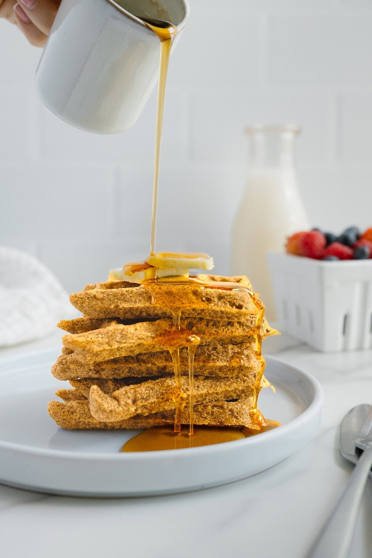 A hand pours syrup from a small pitcher onto a stack of waffles topped with banana slices. In the background, there's a bottle of milk and a container of berries. A fork and knife rest on the plate.