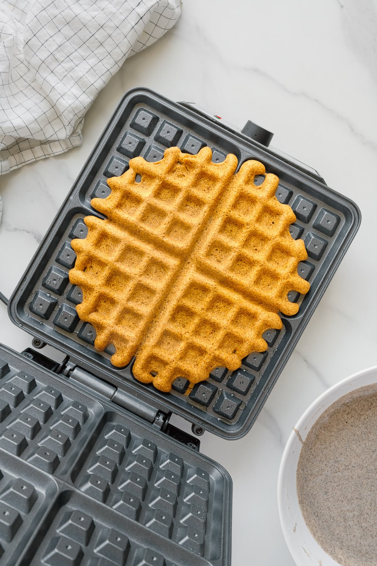 A golden brown buckwheat waffle sits on an open waffle maker, with a white and gray kitchen towel nearby and a bowl of gluten-free batter in the corner on a white countertop.