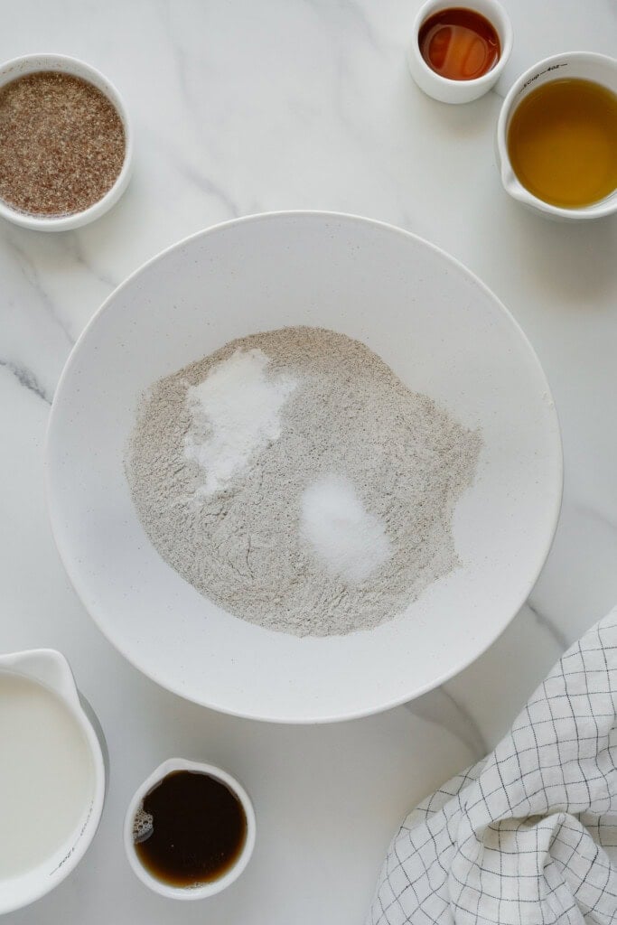 A white bowl filled with flour, baking soda, and salt sits on a marble countertop, surrounded by small bowls containing liquid ingredients, flaxseed meal, and a checkered kitchen towel.