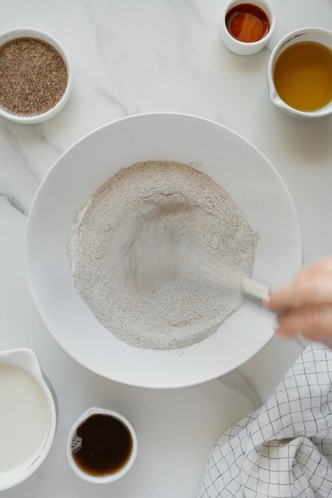 A hand whisks dry ingredients in a white bowl surrounded by small bowls containing liquid ingredients on a marble countertop, with a checkered cloth partially visible nearby.