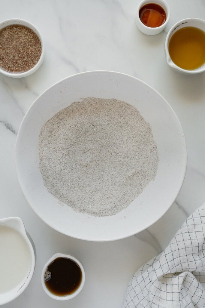 A large white bowl of flour sits on a marble surface, surrounded by small bowls containing milk, maple syrup, vanilla, and another liquid, next to a white checkered cloth.