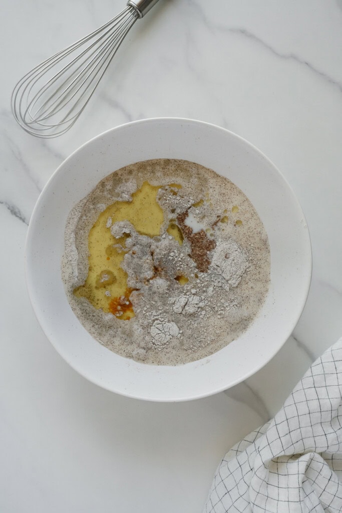 A white bowl containing flour, eggs, and wet ingredients ready to be mixed, sits on a marble surface next to a metal whisk and a white cloth with a black grid pattern.