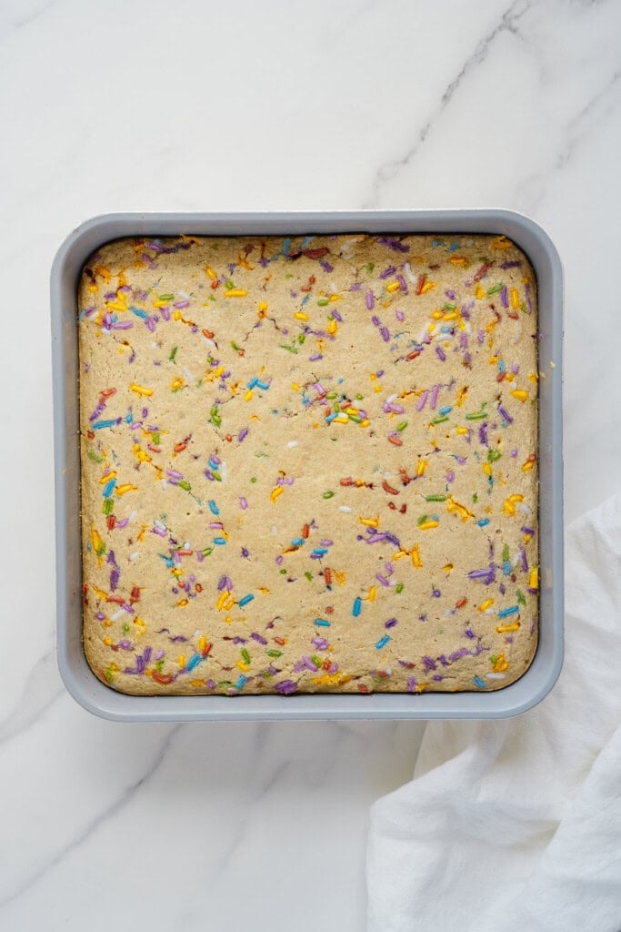 A square baking pan filled with a freshly baked cake topped with colorful sprinkles, resting on a white marble surface with part of a white cloth visible in the corner.