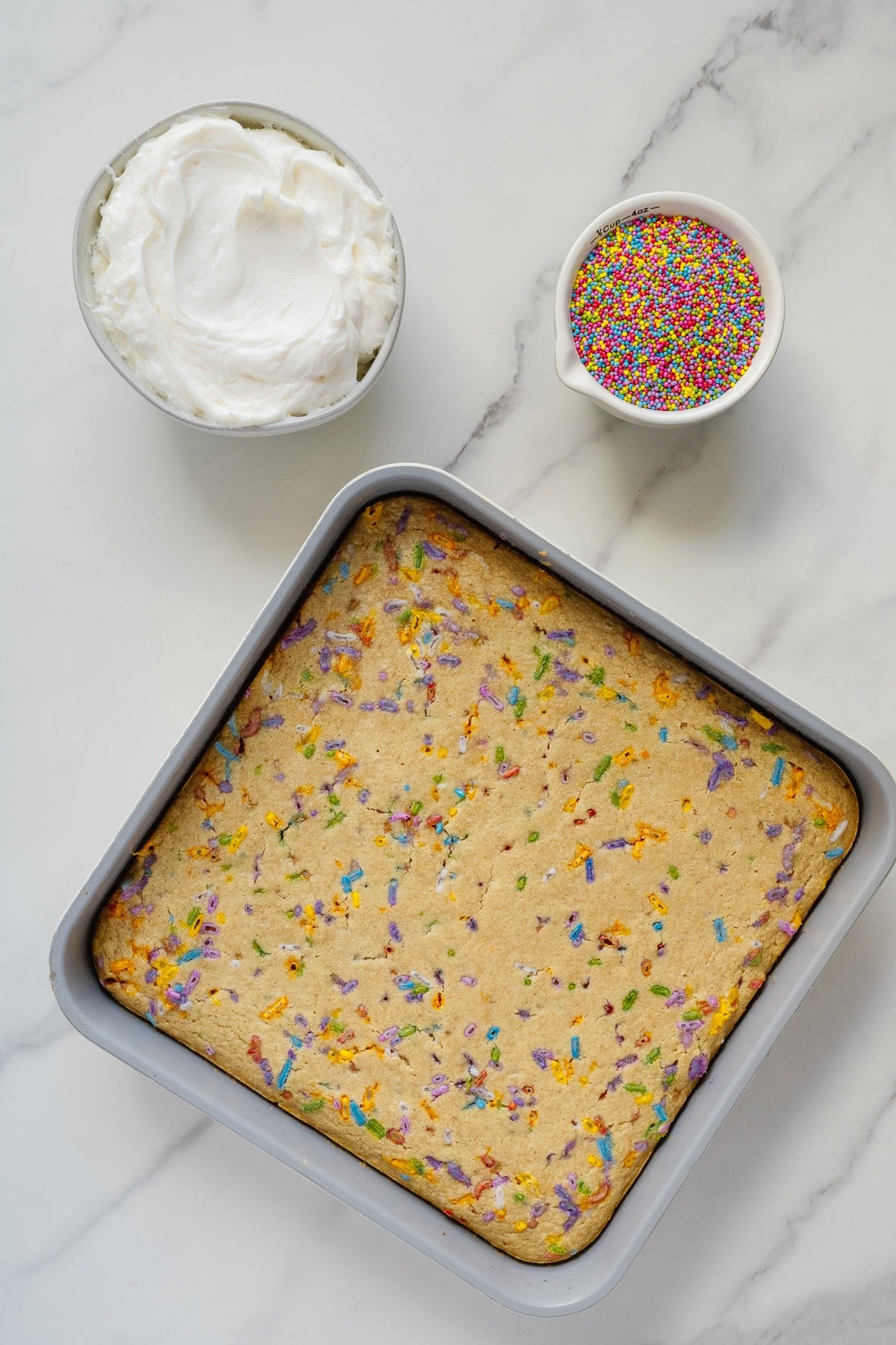A square funfetti cake in a baking pan sits on a marble surface, with a bowl of white frosting and a bowl of colorful sprinkles nearby, ready for decorating.