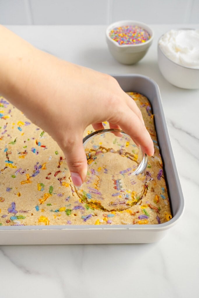 A hand uses a glass cup to cut a circle out of a baked funfetti cake in a rectangular pan. In the background are bowls of colorful sprinkles and whipped cream on a white countertop.