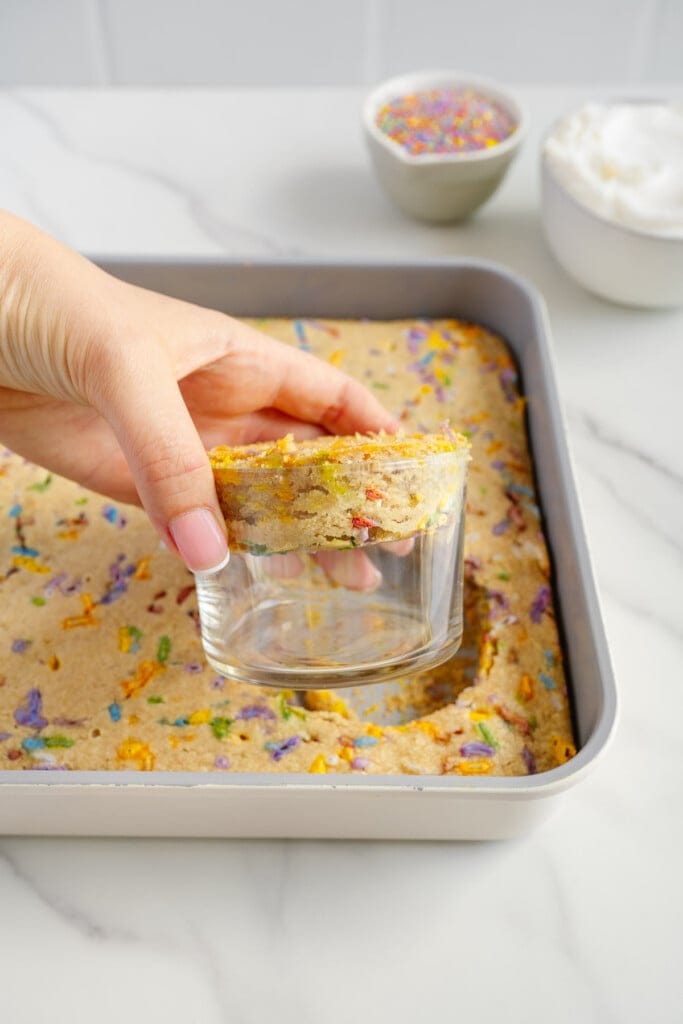 A hand uses a glass to cut a round piece from a rectangular tray of funfetti cake, with bowls of sprinkles and white frosting in the background on a marble surface.