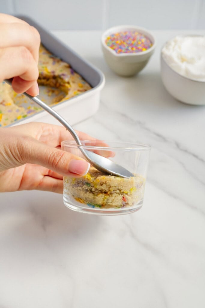 A hand uses a spoon to scoop funfetti cake into a small glass, with a baking tray of cake, a bowl of whipped topping, and a bowl of colorful sprinkles in the background on a white surface.