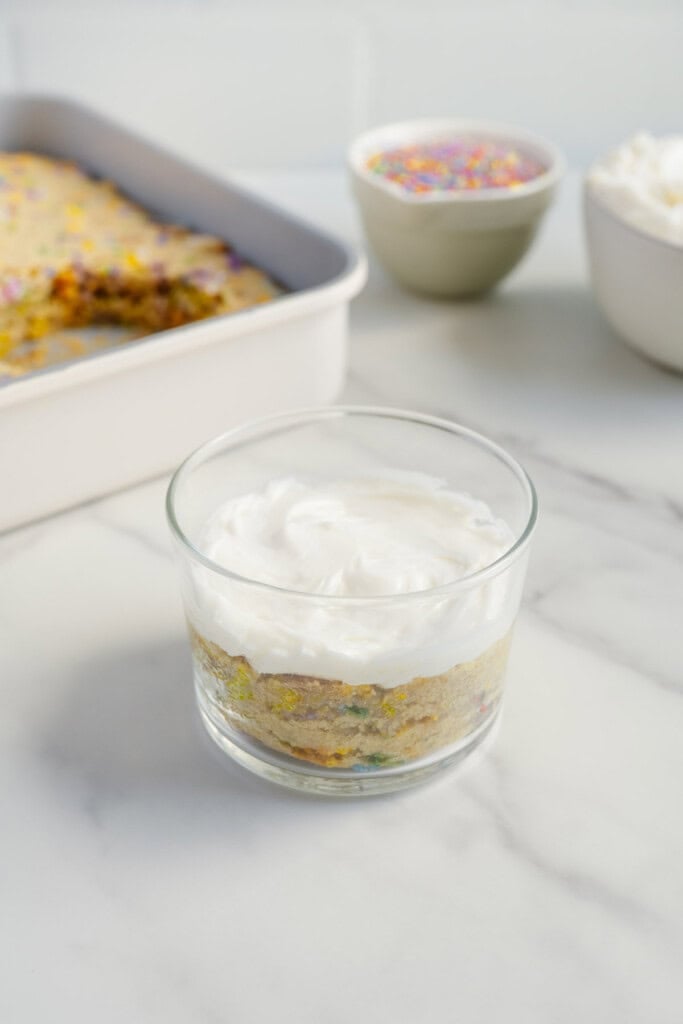 A glass cup filled with layered dessert, featuring a cake-like layer with colorful sprinkles and topped with whipped cream. In the background, a baking dish, a bowl of sprinkles, and a bowl of whipped cream are visible.