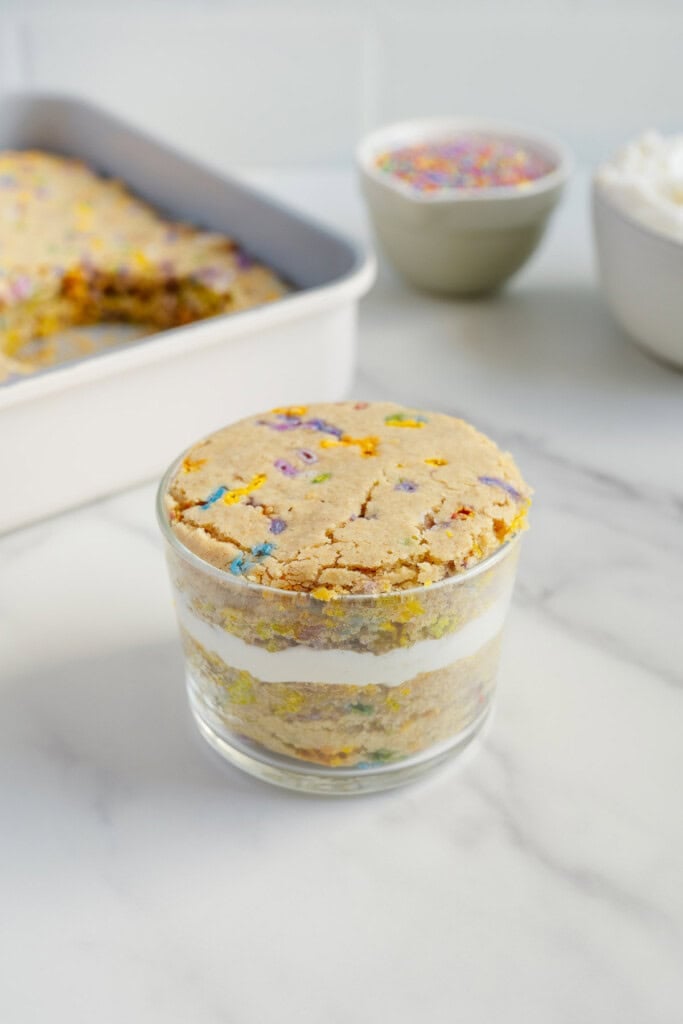A glass filled with layered funfetti cookie crumbles and white cream sits on a marble surface. In the background, a baking pan with more cookie bars and bowls of sprinkles and whipped topping are visible.