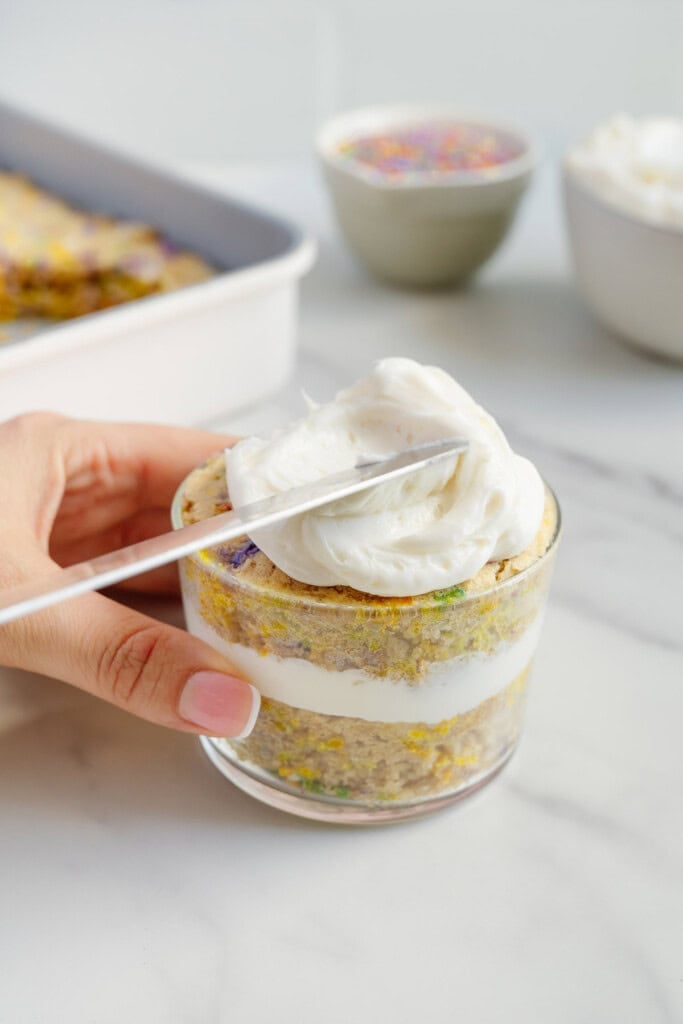A hand holds a glass cup filled with layered cake and cream, while a spoon spreads white frosting on top. In the background, there are bowls with sprinkles and more frosting.