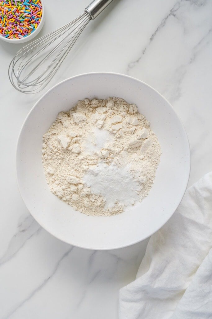 A white bowl filled with flour and baking powder sits on a marble surface. Nearby are a whisk, a small bowl of colorful sprinkles, and a white cloth.