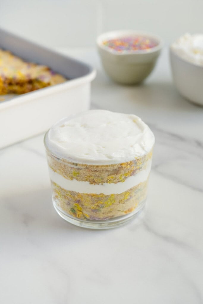A glass jar filled with layers of white frosting and colorful funfetti cake, set on a marble surface. In the background, there's a white baking dish, a bowl of sprinkles, and a bowl of frosting.