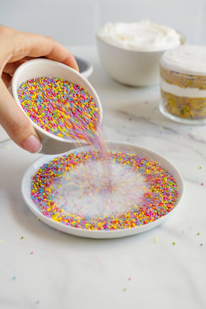 A hand pours colorful round sprinkles from a small bowl onto a shallow dish. In the background, bowls of whipped cream and layered dessert are visible on a white countertop.