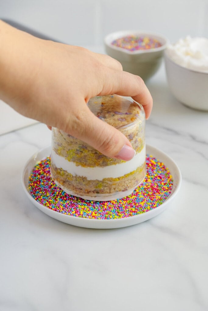 A hand presses a small layered cake with white frosting between plastic molds into a plate of colorful round sprinkles. Bowls of sprinkles and whipped cream are in the background on a marble surface.