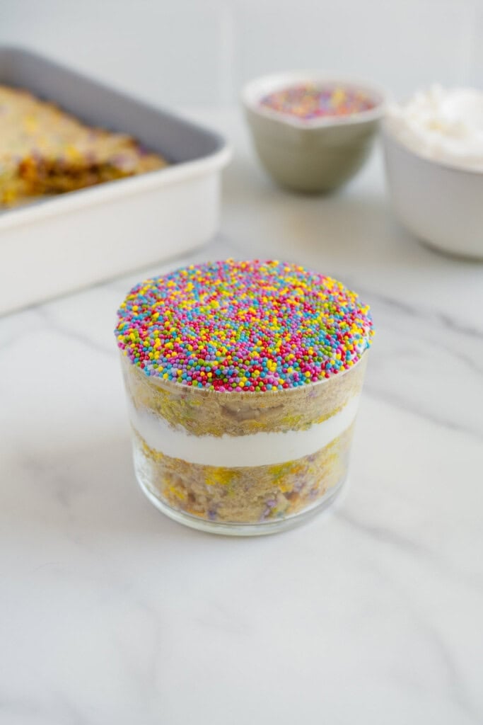 A layered dessert in a glass featuring white cream and cake, topped with colorful round sprinkles. Blurred bowls and a tray with similar dessert are in the background on a white marble surface.