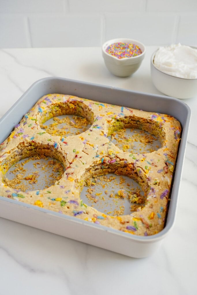 A square baking pan with confetti cake, four large circles cut out, sits on a white surface. In the background are a bowl of rainbow sprinkles and a bowl of white frosting.