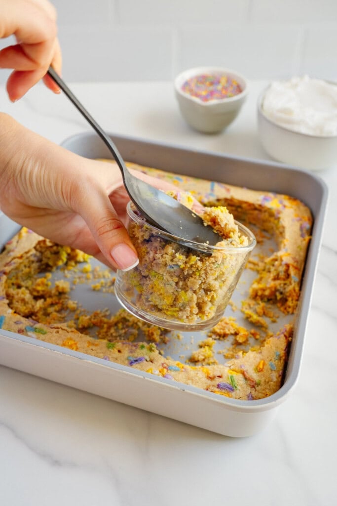 A hand scoops a serving of crumbly cake from a square baking pan into a glass jar using a spoon. In the background, a bowl of whipped topping and a dish of colorful sprinkles sit on a white surface.