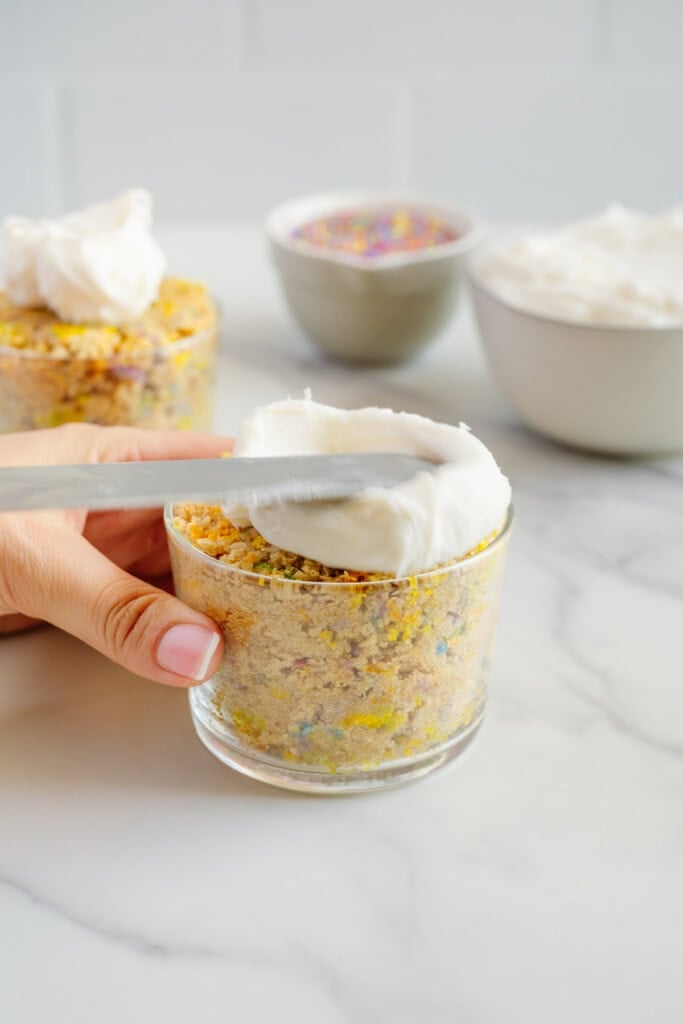 A hand spreads white frosting on top of a small glass filled with crumbled funfetti cake. Bowls of whipped topping and colorful sprinkles are in the background on a white countertop.