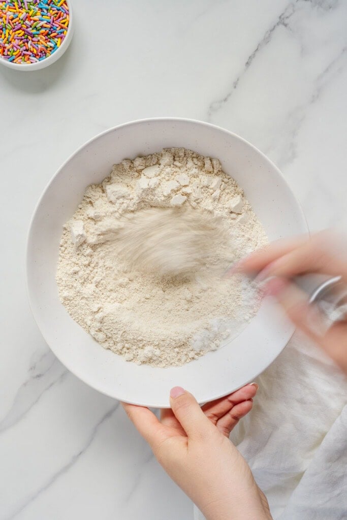 A person stirs flour in a white bowl on a marble surface, with a small bowl of colorful sprinkles nearby.