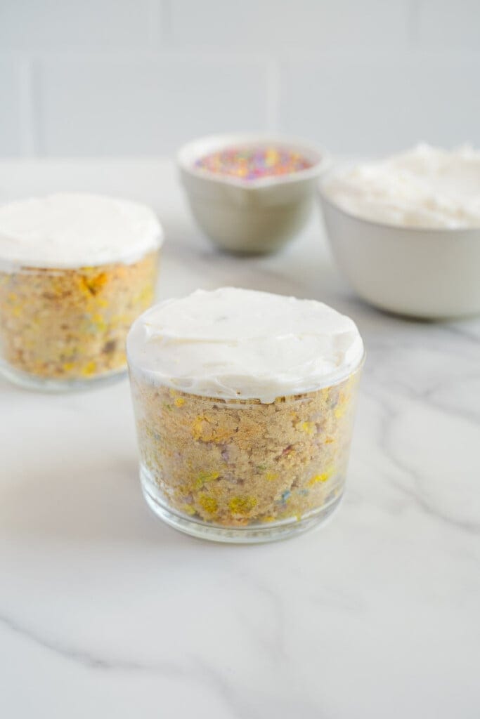 Two small glass jars filled with funfetti cake and topped with white frosting sit on a marble surface. In the background, there are two bowls, one with colorful sprinkles and one with white frosting.