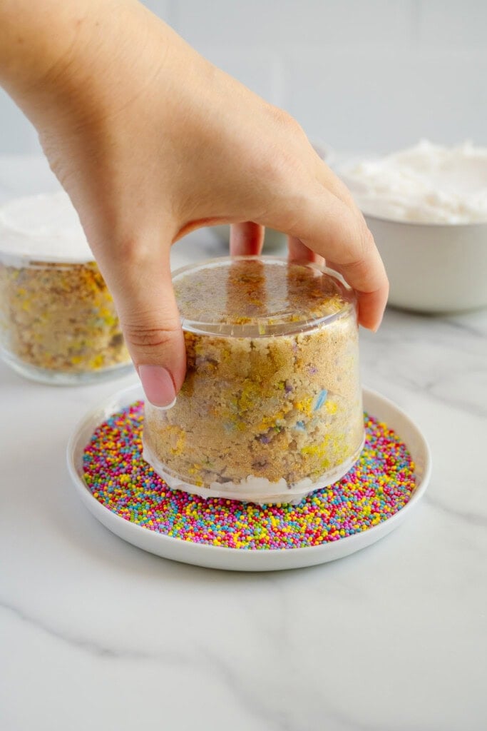 A hand lifts an upside-down jar containing funfetti cake onto a plate filled with colorful round sprinkles. A bowl of whipped cream and another jar of cake are in the background.