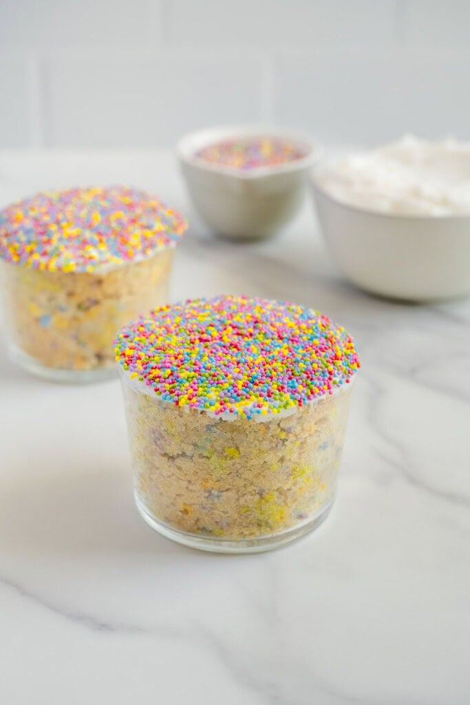 Two small glass cups filled with cake, topped with colorful round sprinkles, sit on a marble countertop. In the background, bowls with more sprinkles and whipped topping are visible.