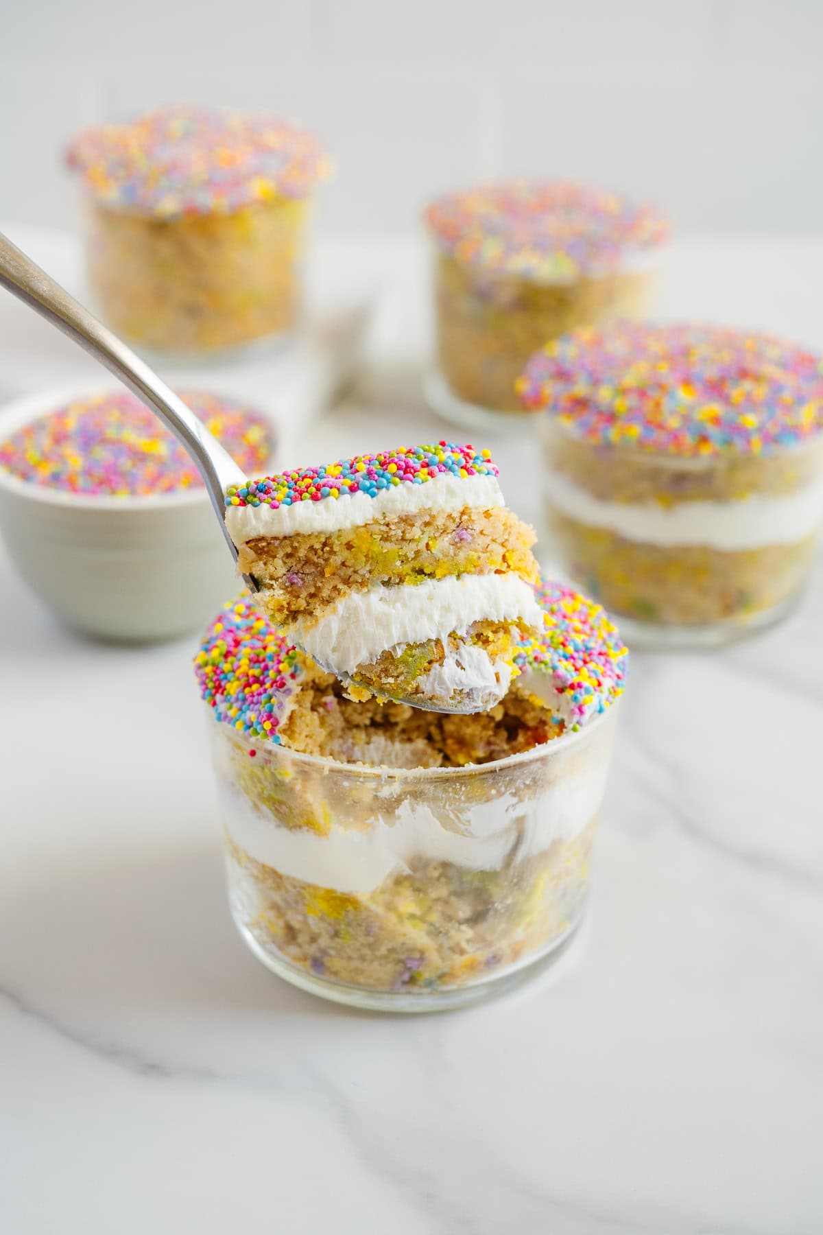 A spoon lifting a layered dessert with cake, white cream, and colorful rainbow sprinkles from a glass jar; more similar desserts are in the background on a white surface.