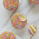 Four round cupcakes covered in colorful sprinkles sit on a white surface. One cupcake has a bite taken out, with a spoon holding a piece of cake and creamy filling beside it.