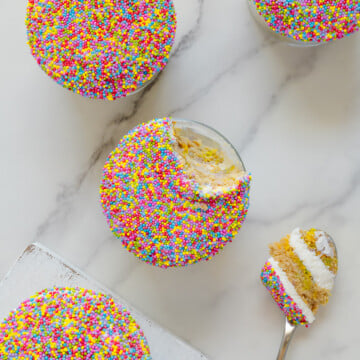 Four round cupcakes covered in colorful sprinkles sit on a white surface. One cupcake has a bite taken out, with a spoon holding a piece of cake and creamy filling beside it.