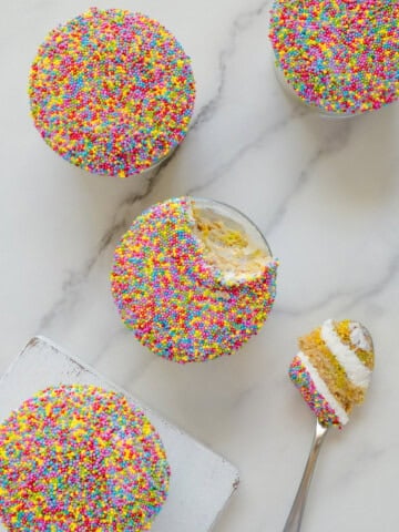Four round cupcakes covered in colorful sprinkles sit on a white surface. One cupcake has a bite taken out, with a spoon holding a piece of cake and creamy filling beside it.