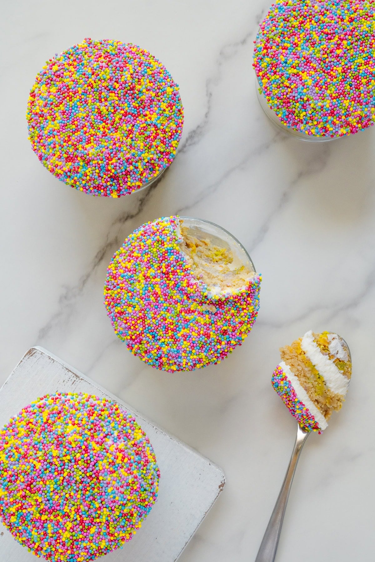 Four round cupcakes covered in colorful sprinkles sit on a white surface. One cupcake has a bite taken out, with a spoon holding a piece of cake and creamy filling beside it.