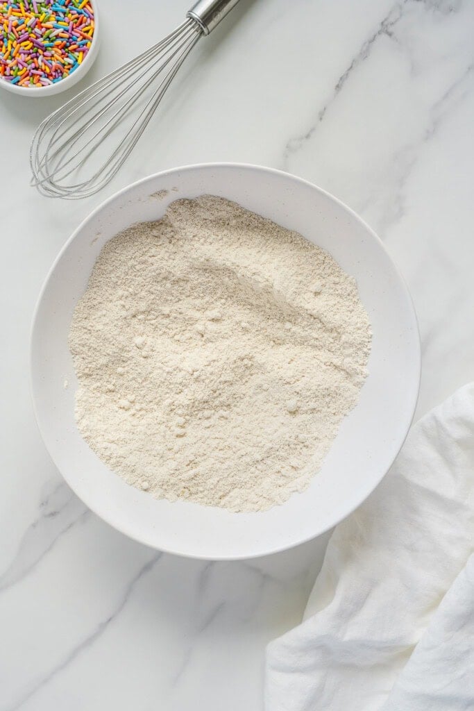 A large white bowl filled with flour sits on a marble countertop. Nearby are a metal whisk, a small bowl of colorful sprinkles, and part of a white cloth.